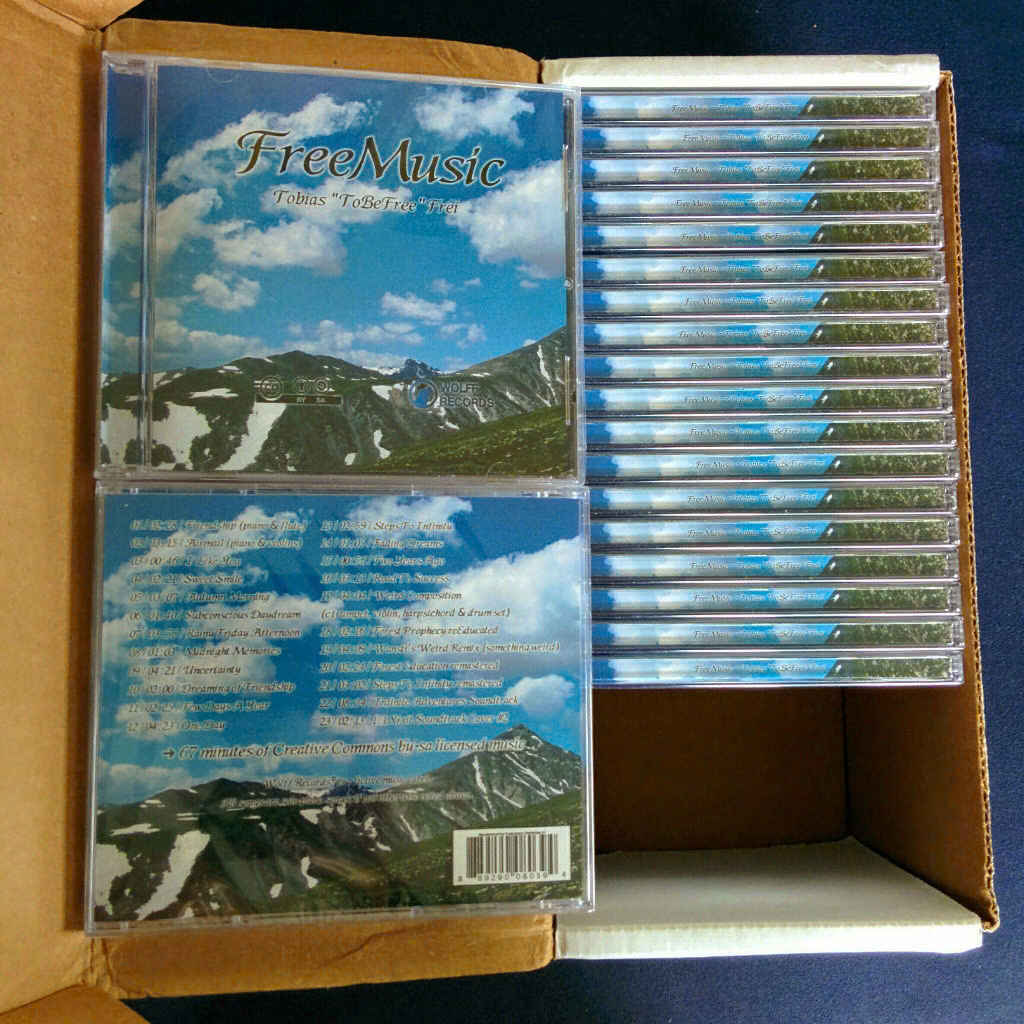 Ein von oben fotografiertes Paket mit 20 FreeMusic-CD-Hüllen, die bis auf zwei Ausnahmen mit ihrer schmalsten Seite (den Rücken) nach oben zum Betrachter zeigen. Die beiden Ausnahmen sind zwei innen auf den aufgeklappten Paketdeckel gelegte Exemplare, die Vorder- und Rückseite der CD-Hüllen präsentieren. Auch hier ist wieder das Alpen-Motiv sichtbar; die Rückseite enthält eine Liste der Lieder, die auf der CD enthalten sind.